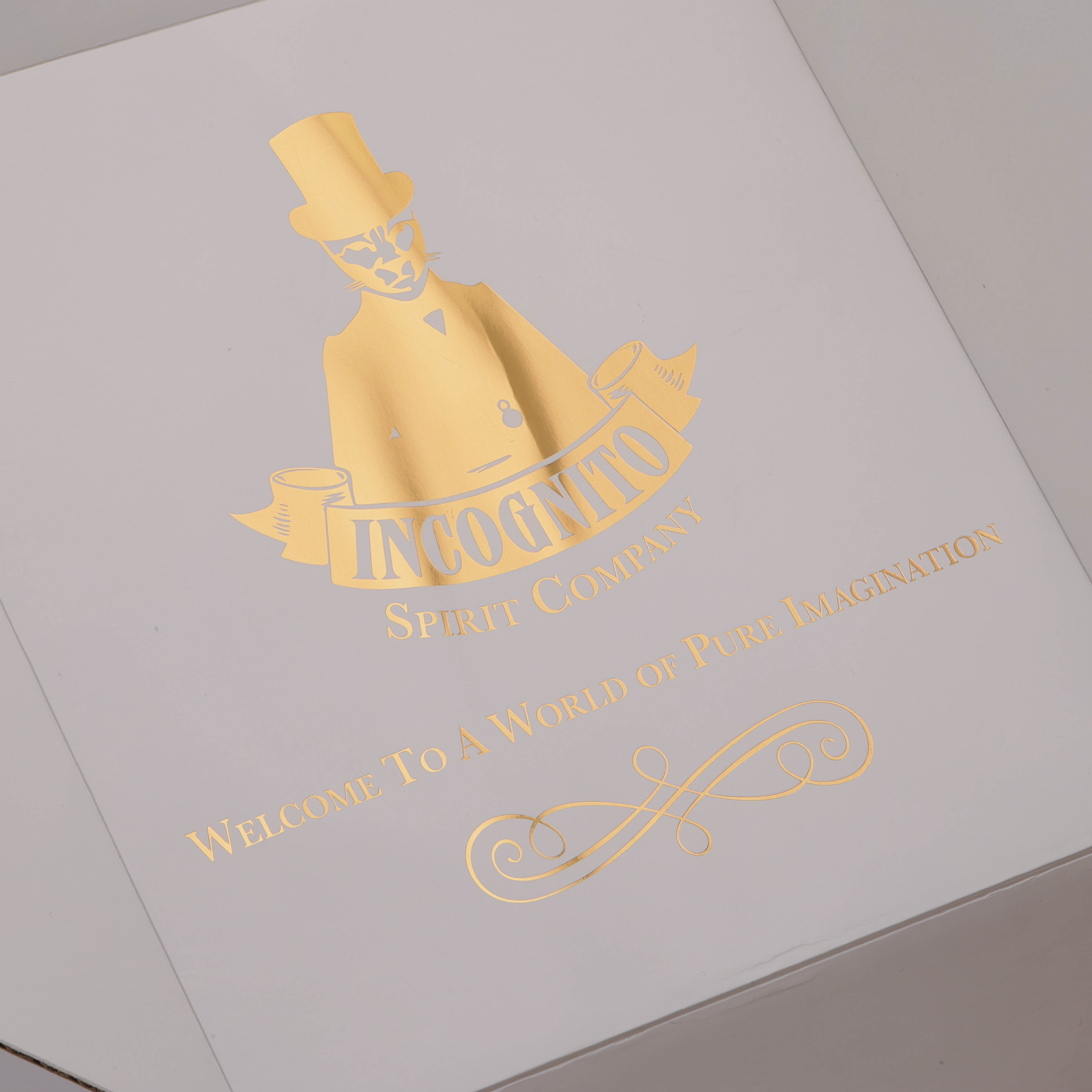 A close-up of a white box lid featuring elegant gold foil branding for 'Incoonto Spirit Company' with a stylized figure in a top hat and the text 'Welcome To A World of Pure Imagination'.