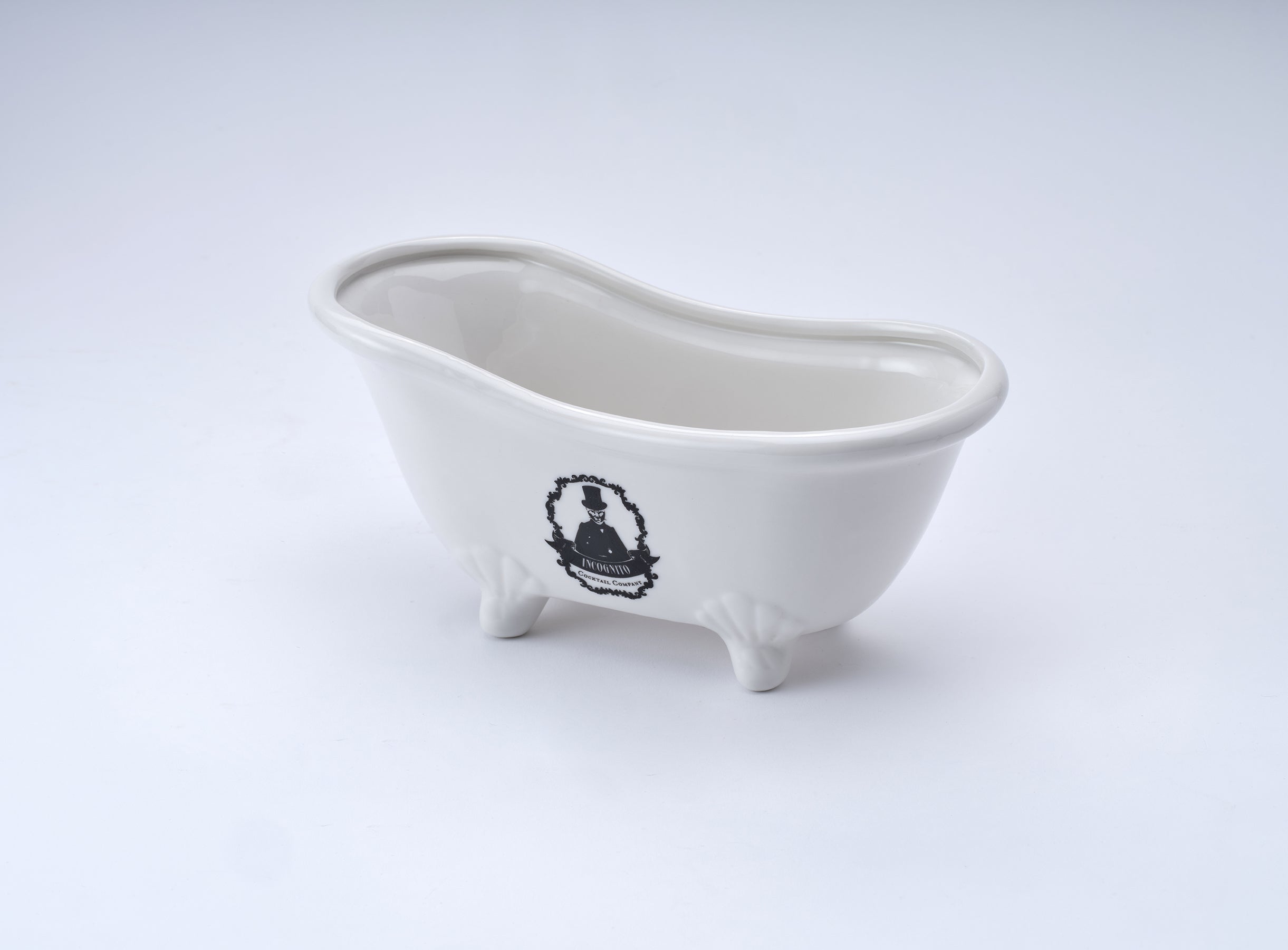 A small white ceramic novelty bathtub figurine with a black crest emblem on the side, set against a seamless light gray background.