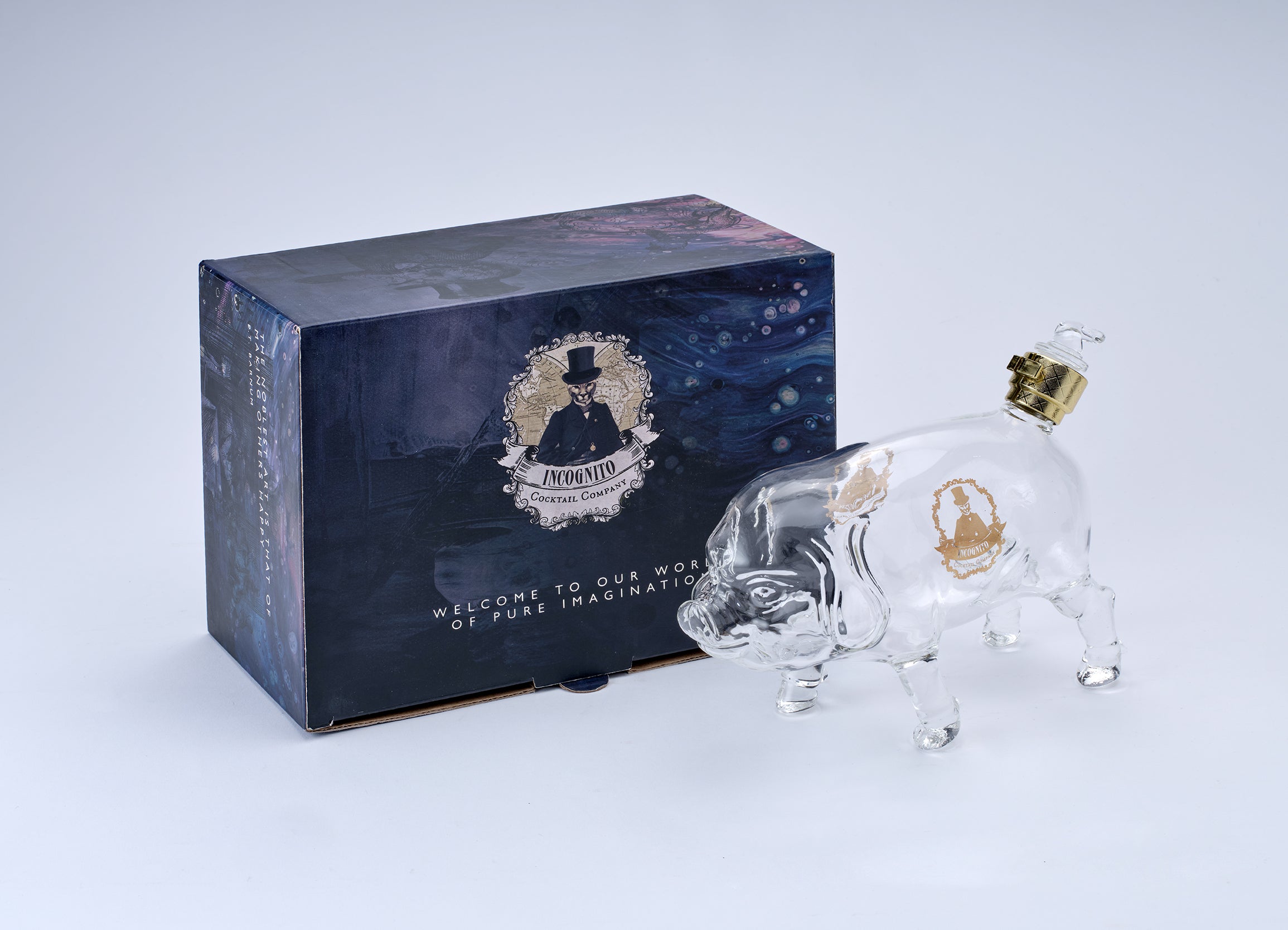 A clear glass pig-shaped bottle with a gold cap and gold emblem, displayed next to its dark blue illustrated packaging box on a light gray background.