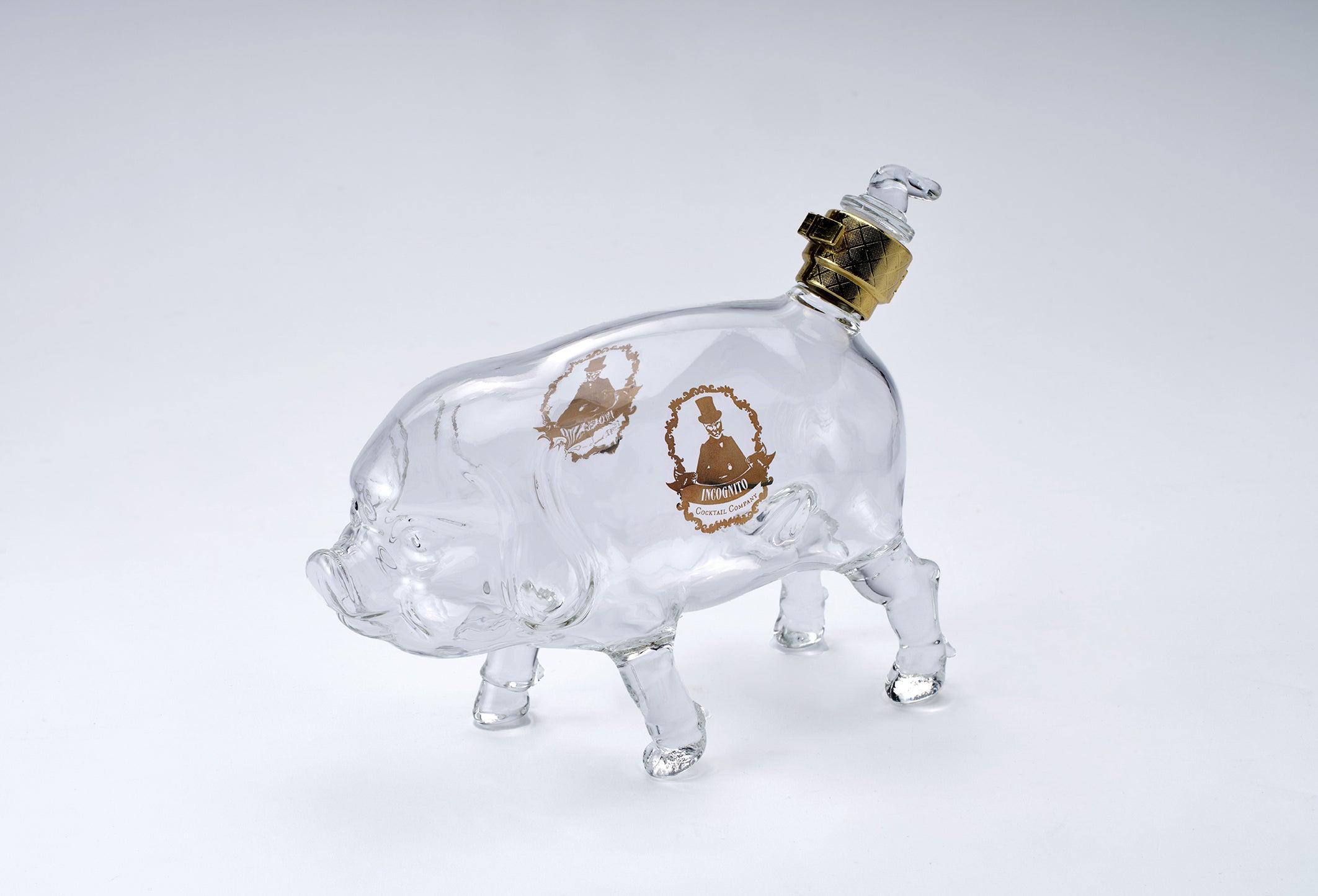A clear glass pig-shaped bottle with gold crest engravings and a gold-toned metal cap, photographed on a seamless light gray background.