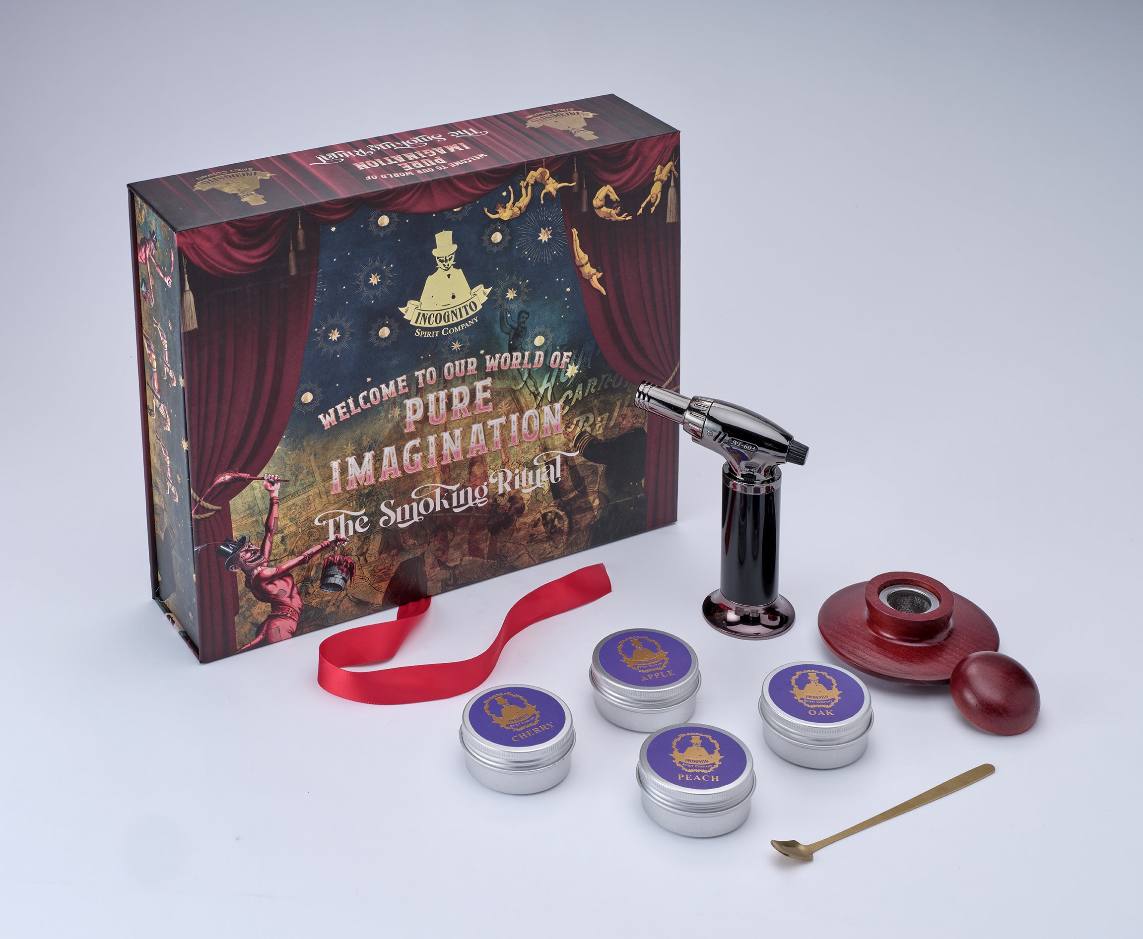 A themed smoking accessory set displayed with its retail box on a light gray studio background. The set includes a black and silver torch lighter, a red mouthpiece, a red ribbon, five small round tin containers with purple lids, a small gold spoon, and a decorative box printed with theatrical-style artwork and the text 'Welcome to our world of Piped Imagination' and 'The Smoking Room.