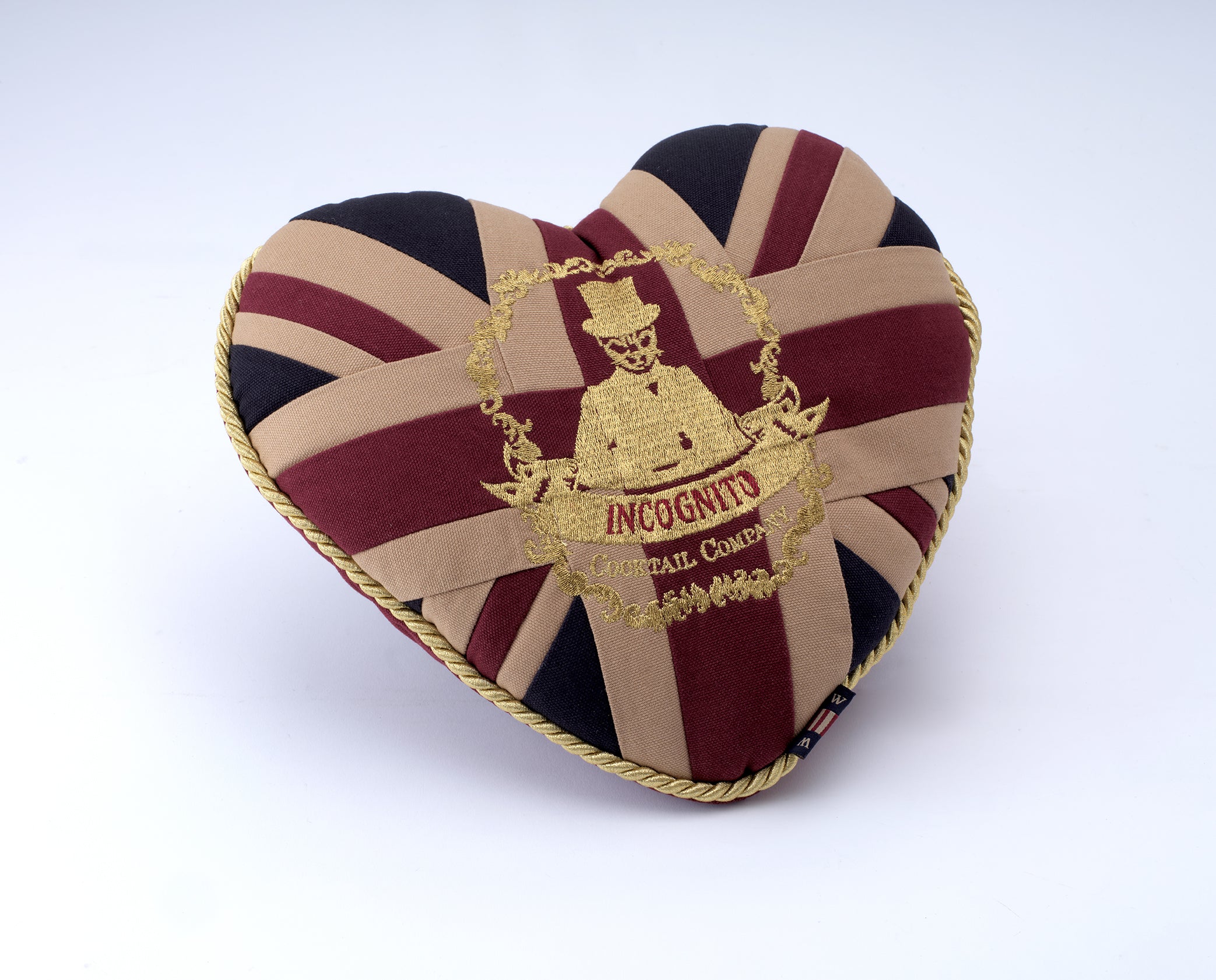 A heart-shaped novelty pillow featuring a Union Jack pattern in beige, burgundy, and black, with a gold embroidered crest in the center reading 'INCognito Your Rail Coach' and a small Union Jack flag at the bottom right corner.