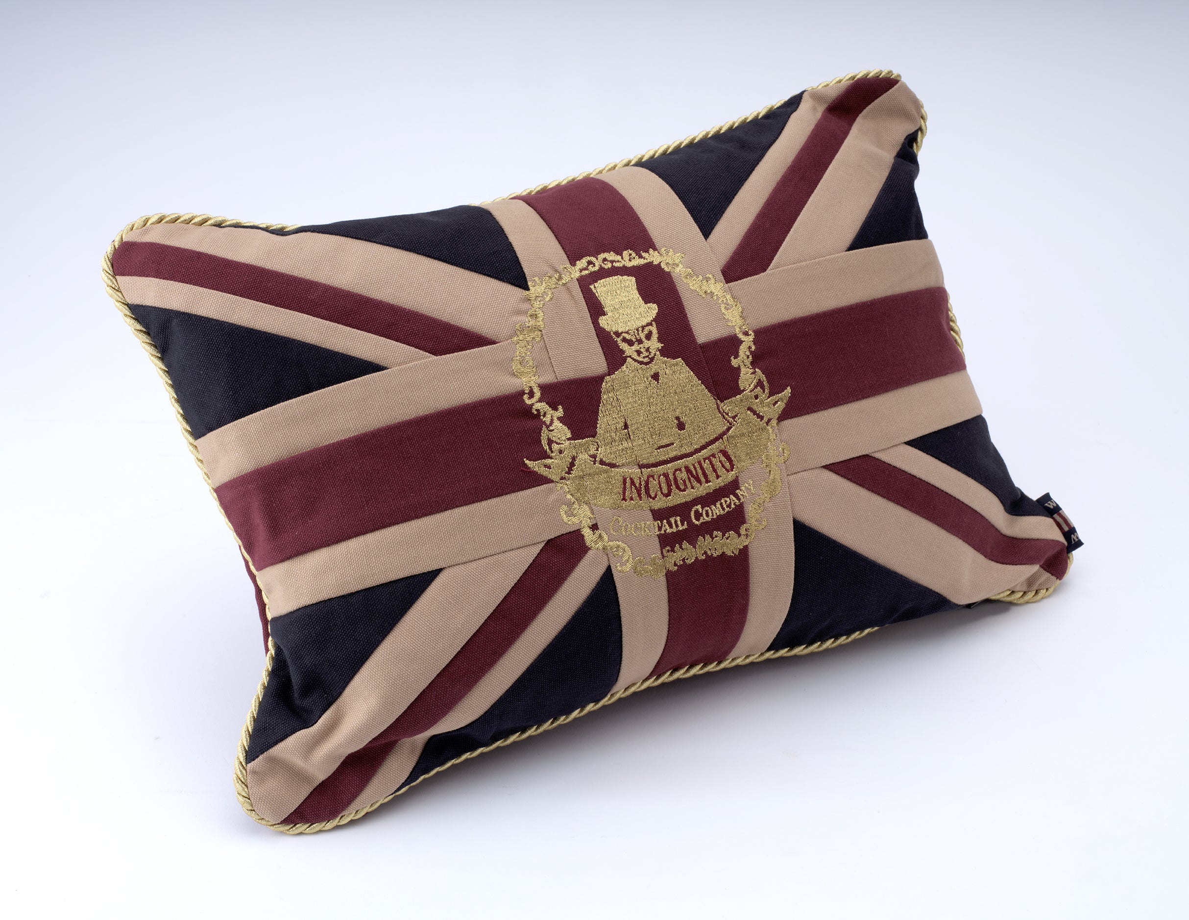 A rectangular decorative pillow featuring a Union Jack flag design in navy blue, beige, and burgundy, with a gold embroidered crest in the center reading 'JACK CUNNITO' and 'Sir, Commando'. The pillow is finished with gold braided trim and is photographed on a clean white background.