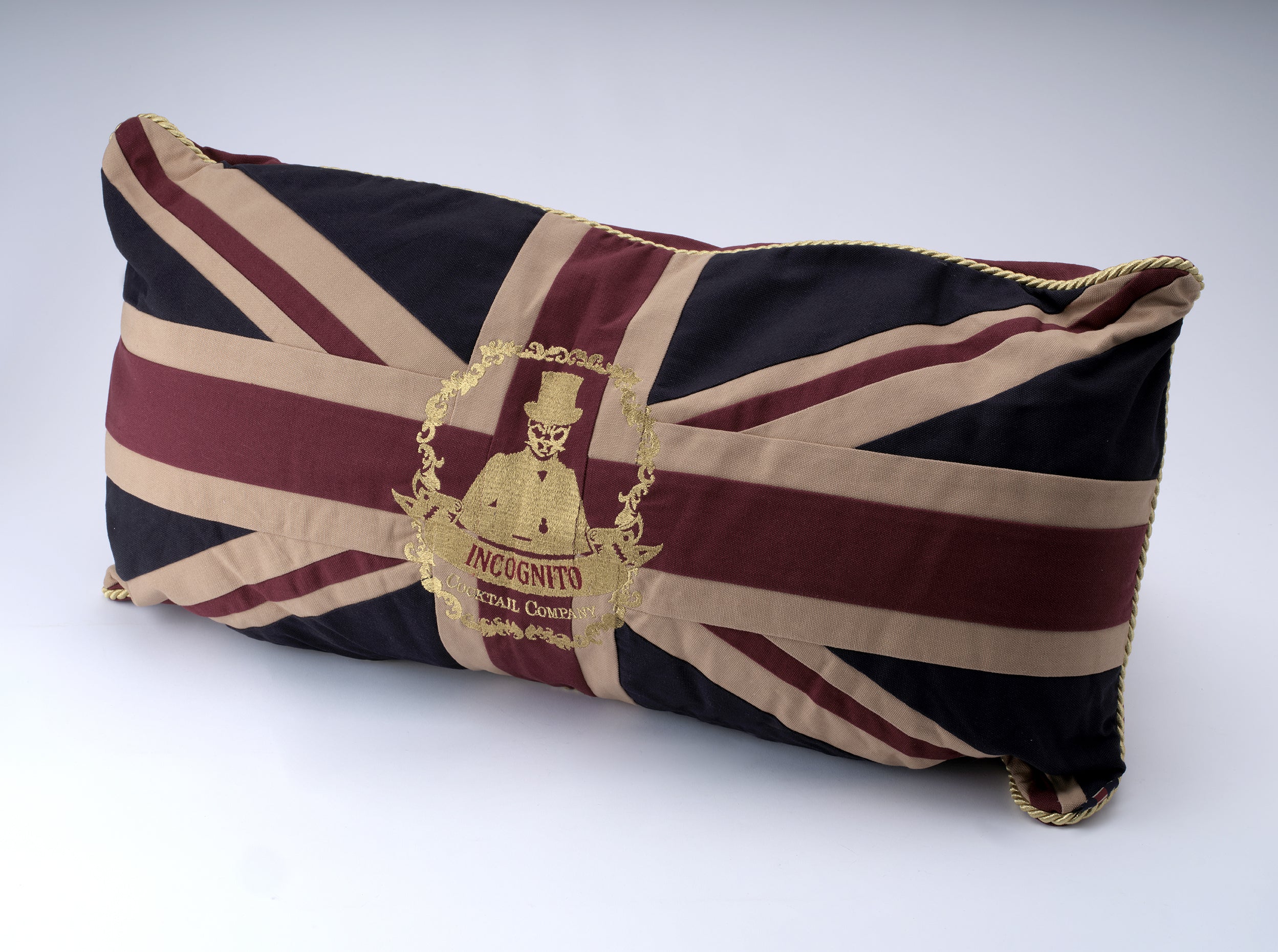 A rectangular novelty pillow featuring a Union Jack flag design in navy blue, burgundy, and beige, with a gold embroidered crest in the center reading 'INCIGNITO'.