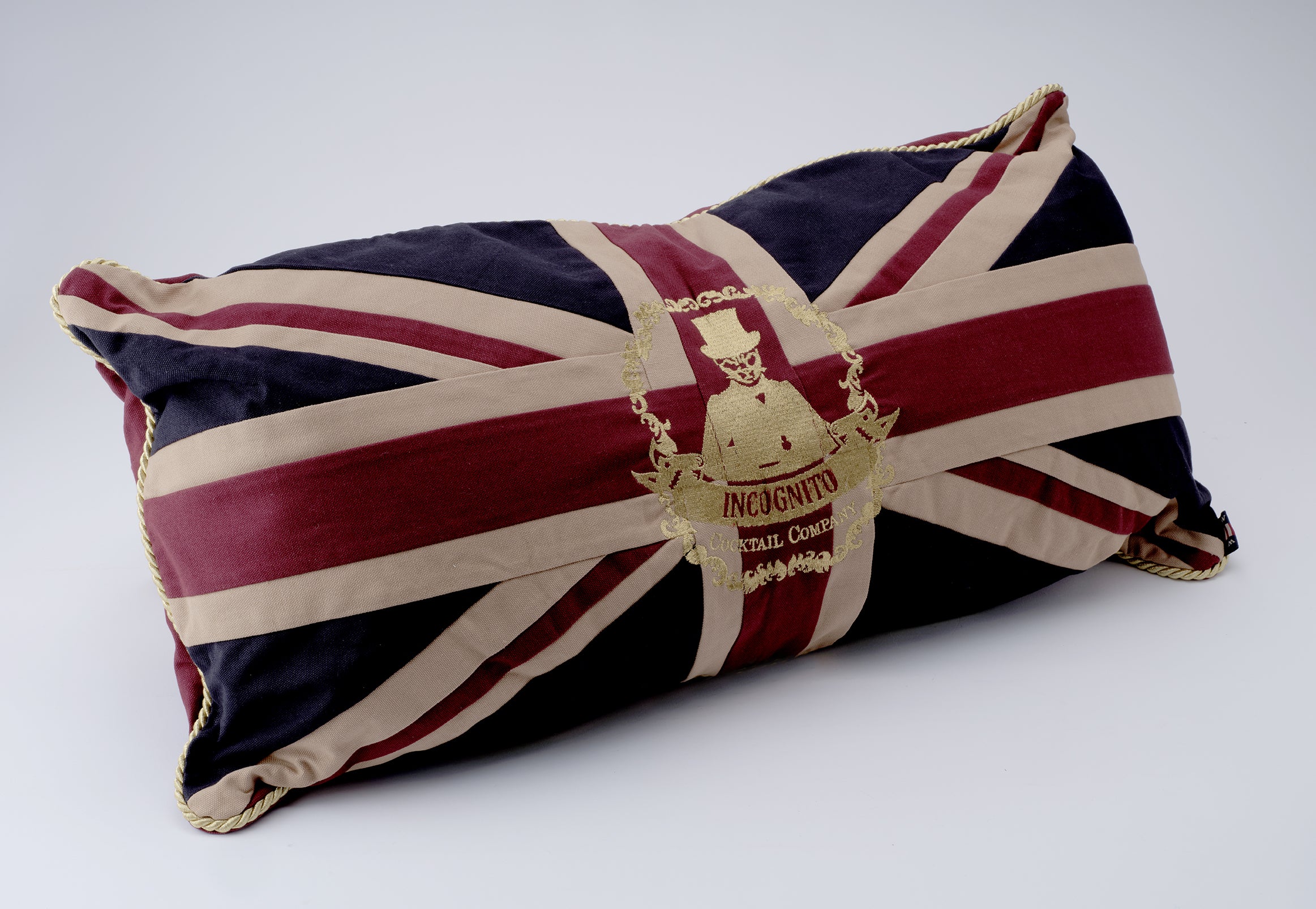 A rectangular throw pillow featuring a Union Jack flag design with a gold embroidered crest in the center, photographed on a light gray background.