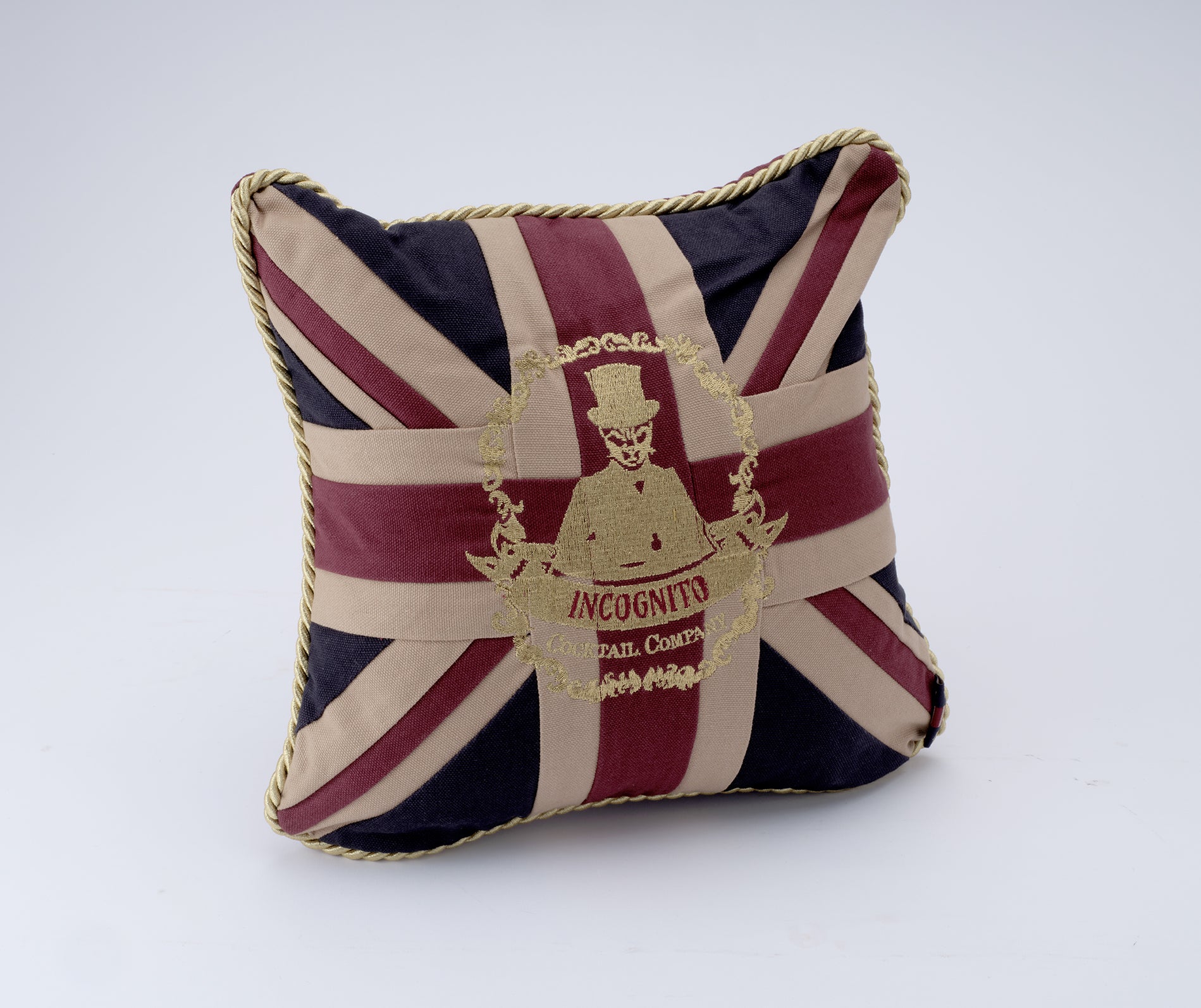 A square decorative throw pillow featuring a Union Jack flag design in burgundy, cream, and navy blue, with a gold embroidered 'Incoognito' character emblem in the center and gold rope trim along the edges.