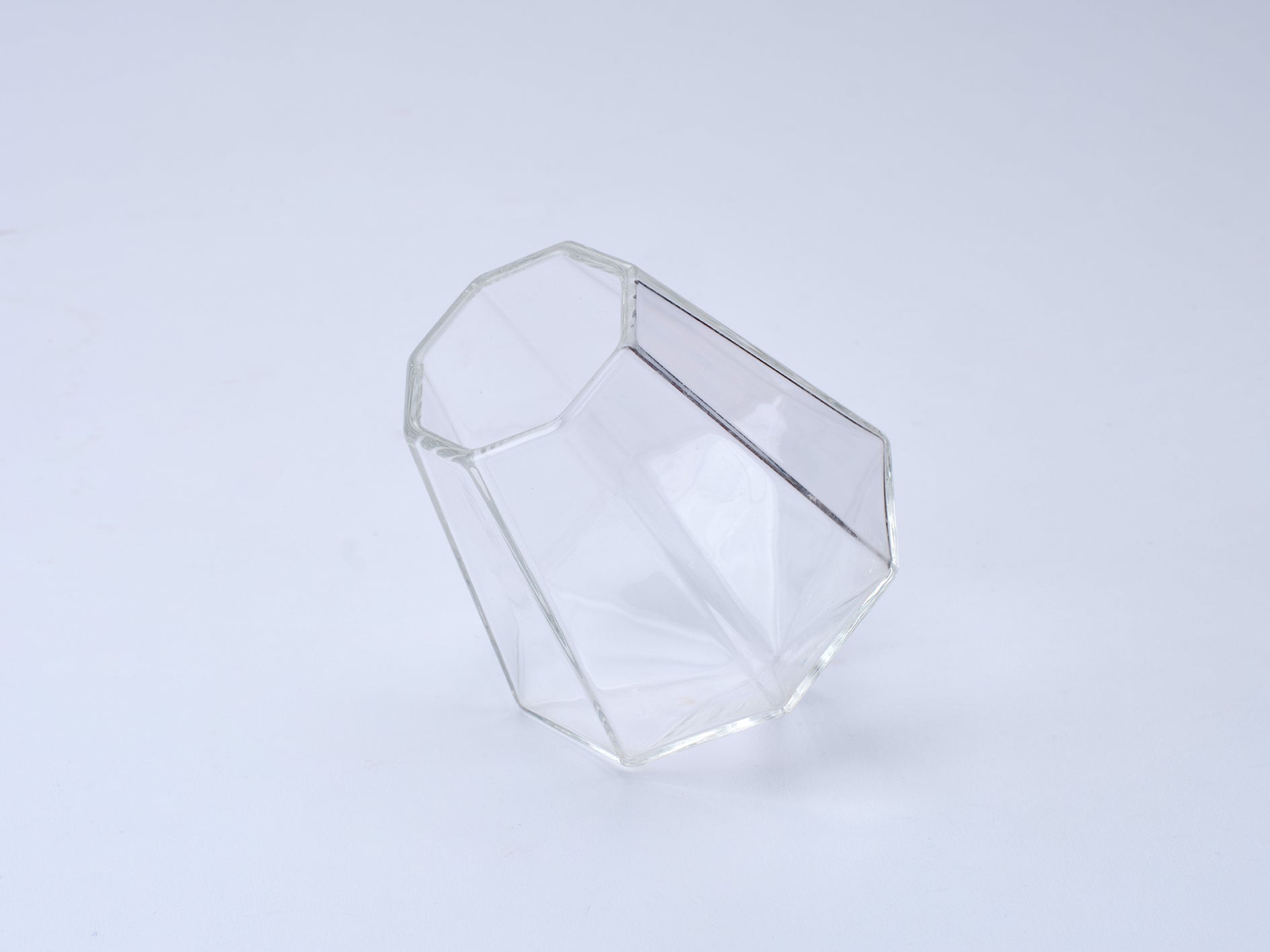 A clear faceted geometric glass container with a diamond-like shape, photographed on a seamless white background.