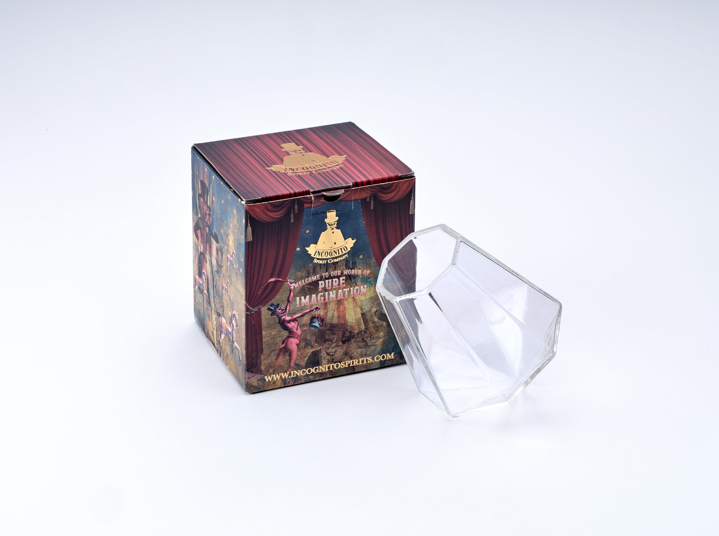 A clear faceted glass tumbler next to its illustrated retail box on a clean white background. The box features a dark red curtain design with gold and multicolor theatrical illustrations and text including 'Theatrical Glasses' and 'Pole Position'.