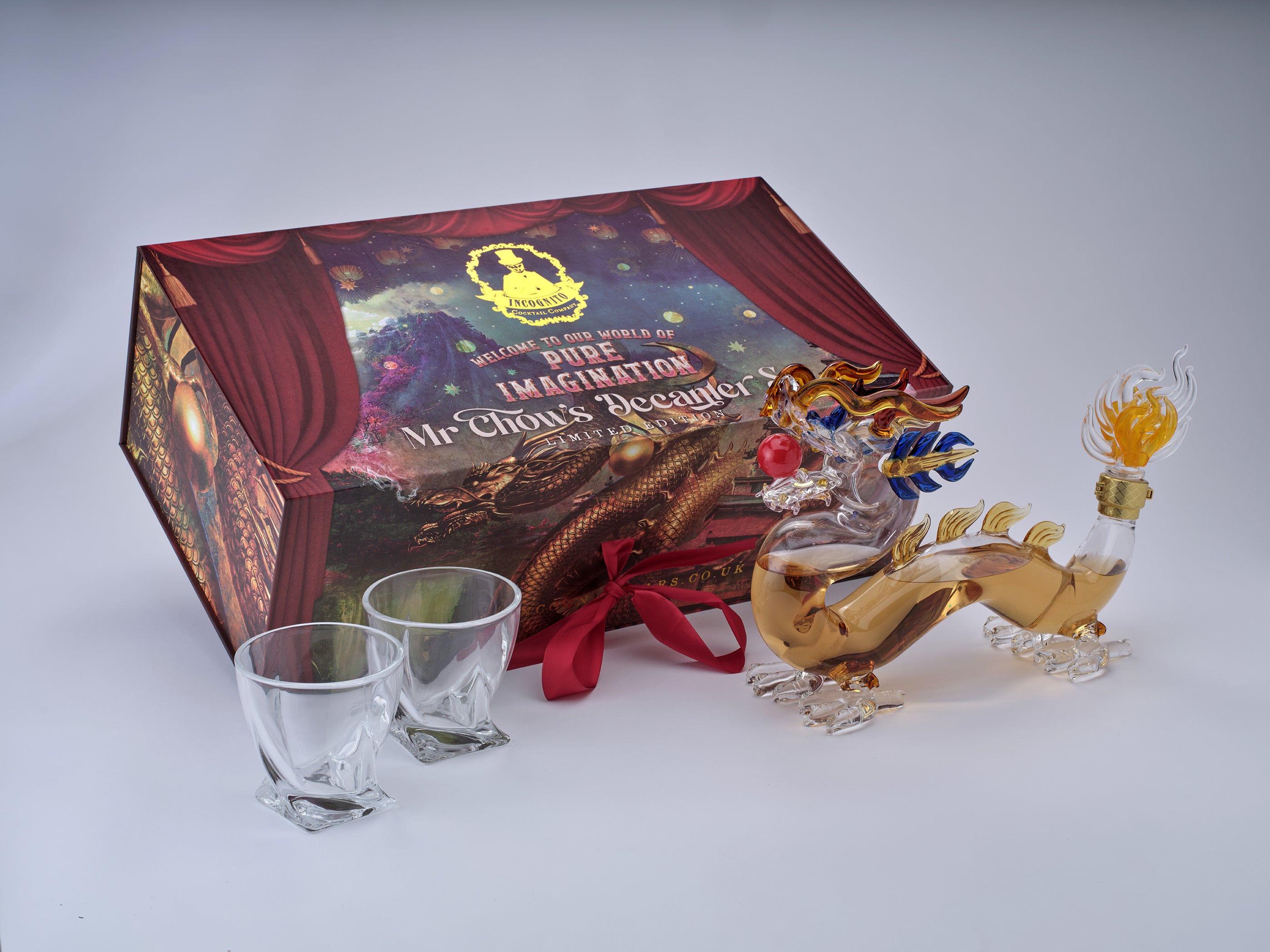 A boxed glass dragon figurine with two small clear glasses placed in front, photographed on a light gray studio background. The box is printed with theatrical-style artwork and the text 'Mr.