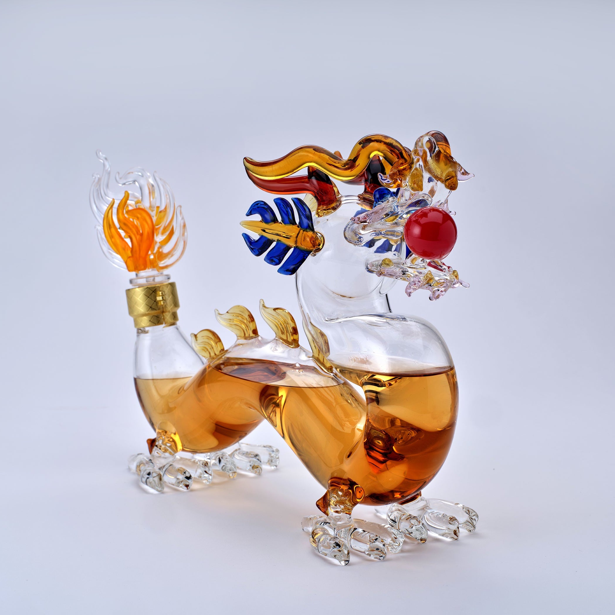 A decorative clear glass dragon figurine filled with amber-colored liquid, featuring gold, blue, red, and orange accents, photographed on a clean light gray background.