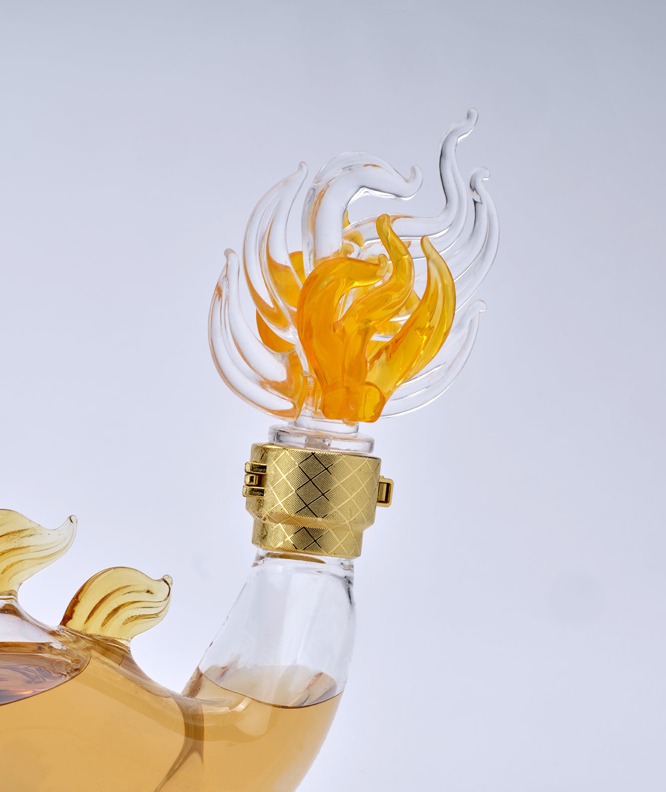 A close-up of a clear glass bottle with a sculptural flame-shaped pourer in amber and transparent glass, topped with a textured gold cap, set against a light gray background.