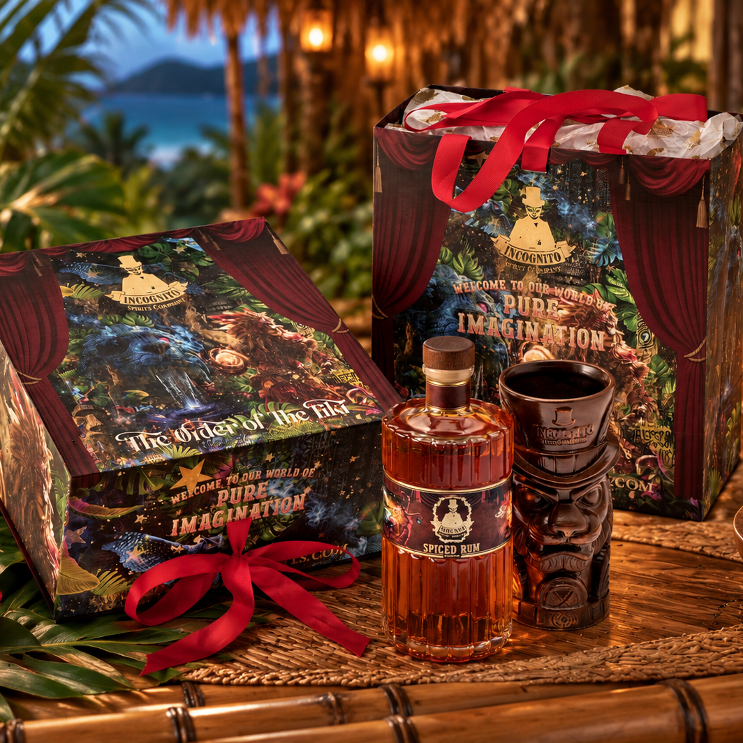 A themed spiced rum gift set displayed on a bamboo table in a tropical bar or lounge setting. The set includes a bottle od Incognito spiced rum, 'illustrated box and a matching tiki-style cup, and a red ribbon, surrounded by props like a tiki mask, pineapple, and lit lanterns.