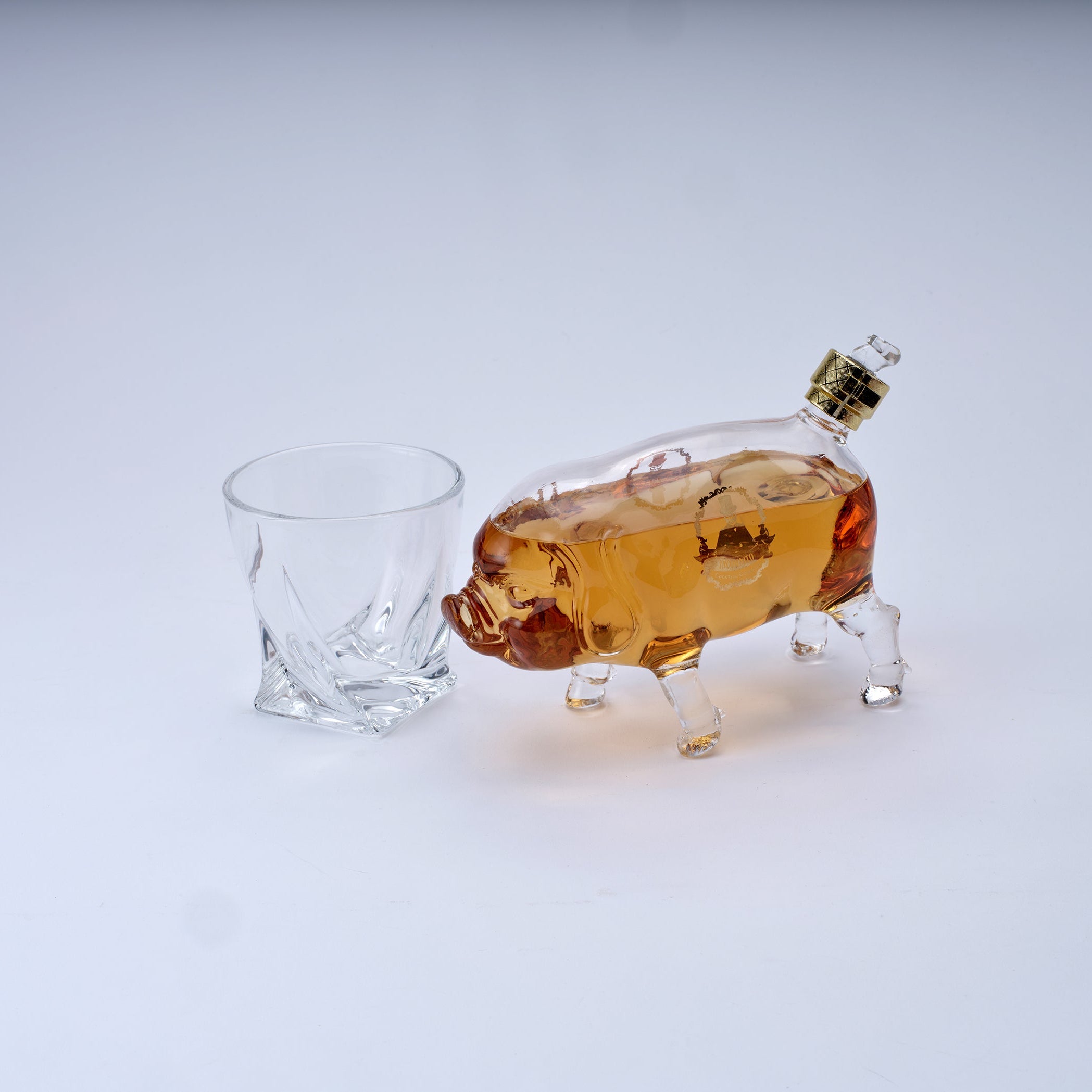A clear glass pig-shaped bottle filled with amber liquor and a small clear glass tumbler, photographed on a clean white studio background.