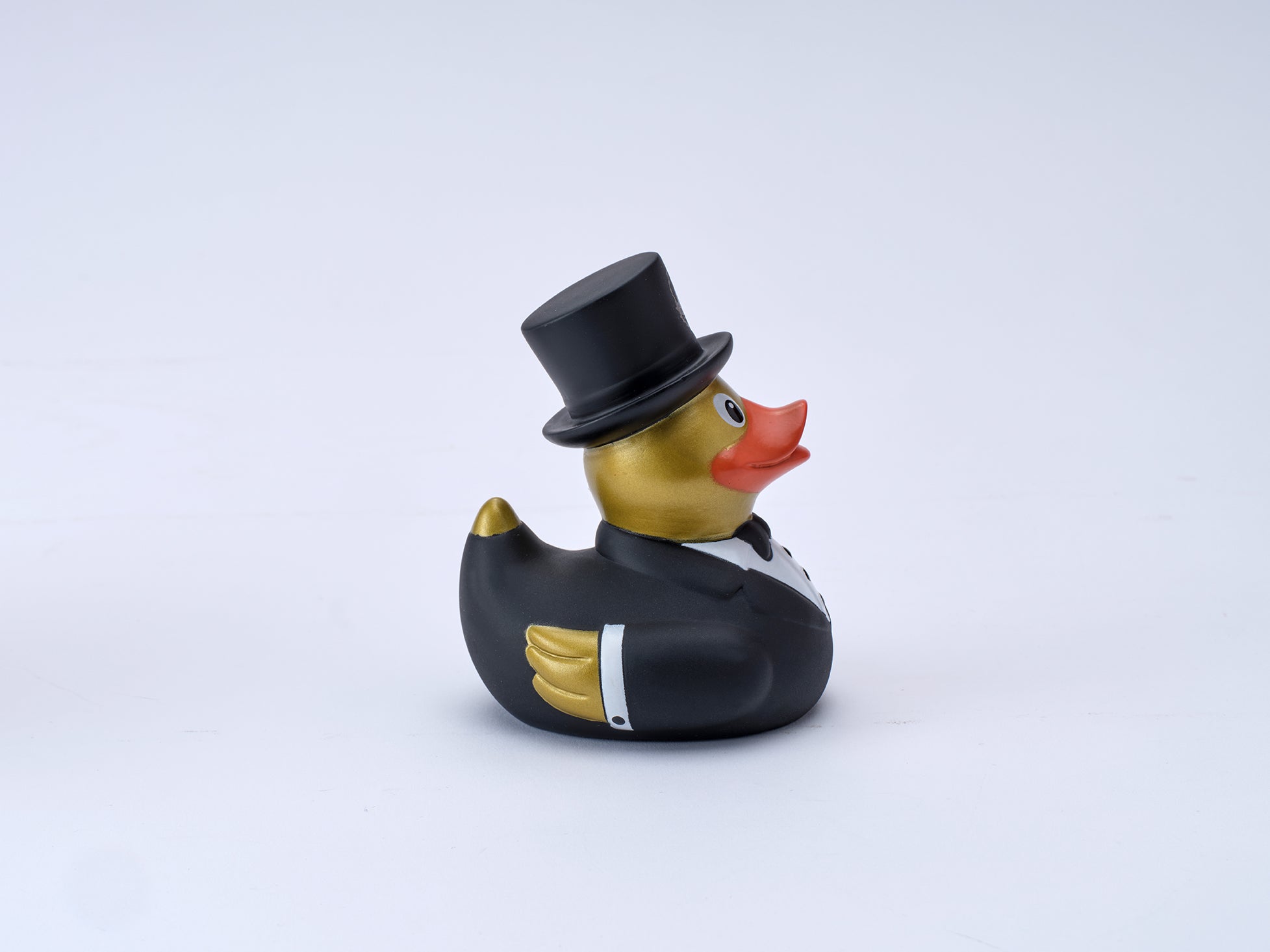 A rubber duck styled as a formal figure, wearing a black top hat, black tuxedo jacket, and white shirt, with a gold body and orange beak, set against a plain light gray background.