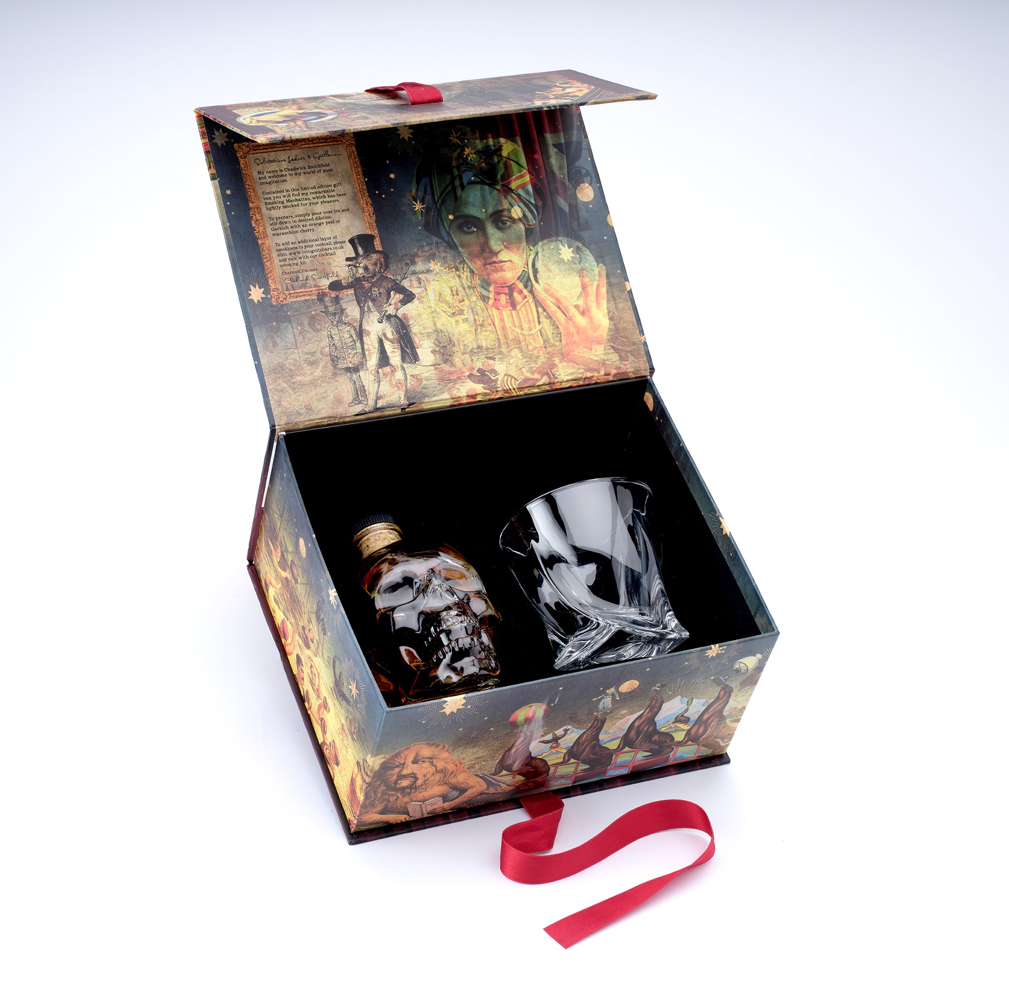 A decorative gift box with an illustrated vintage-style print, shown open on a white background. Inside are a small bottle with a metallic skull cap and a clear glass tumbler.