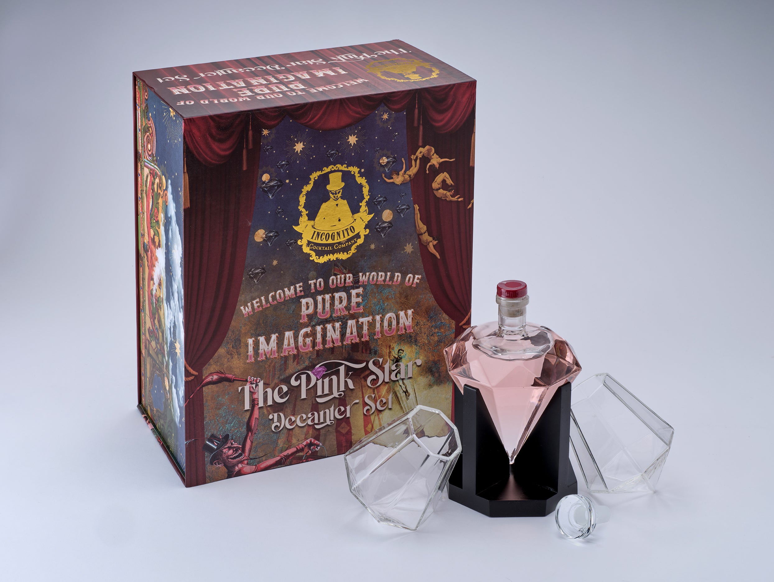 A boxed cocktail set displayed on a light gray studio background. The set includes a decorative pink crystal-shaped cocktail bottle with a black base, two clear faceted tumblers, and a printed presentation box reading 'The Pink Star Cocktail Set' with circus-style artwork.