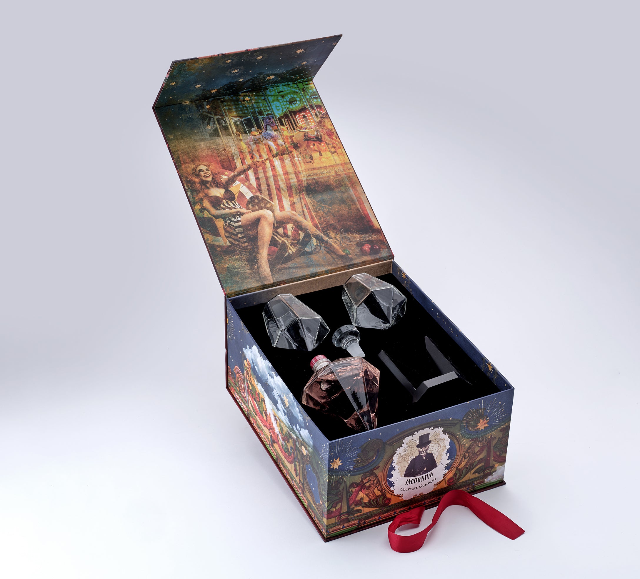An open decorative gift box with illustrated artwork on the exterior and interior, containing a clear glass decanter and two faceted crystal-like glasses, set against a light gray studio background.