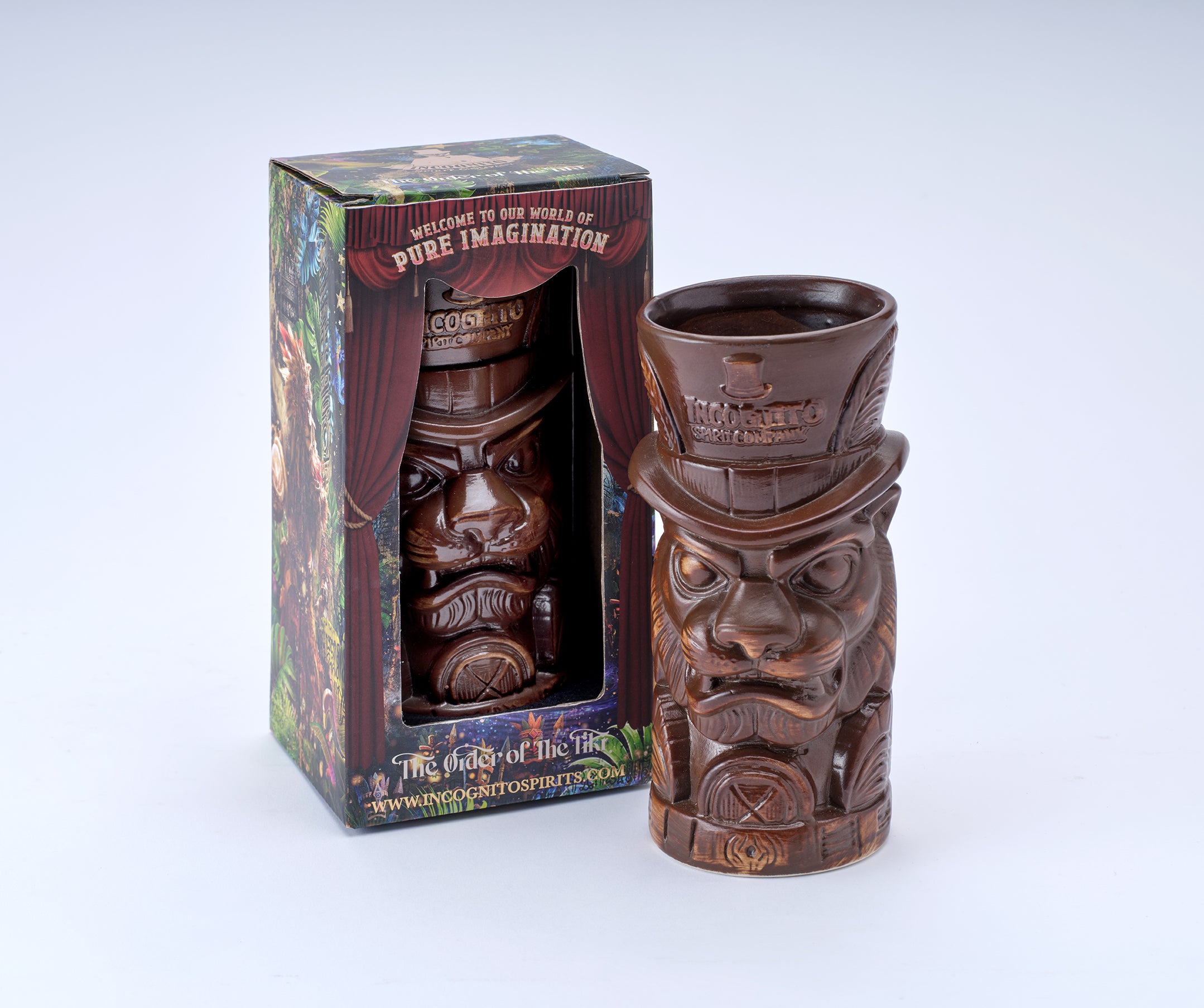 A carved brown tiki-style mug with a face and top hat, shown next to its illustrated retail box on a light gray studio background.
