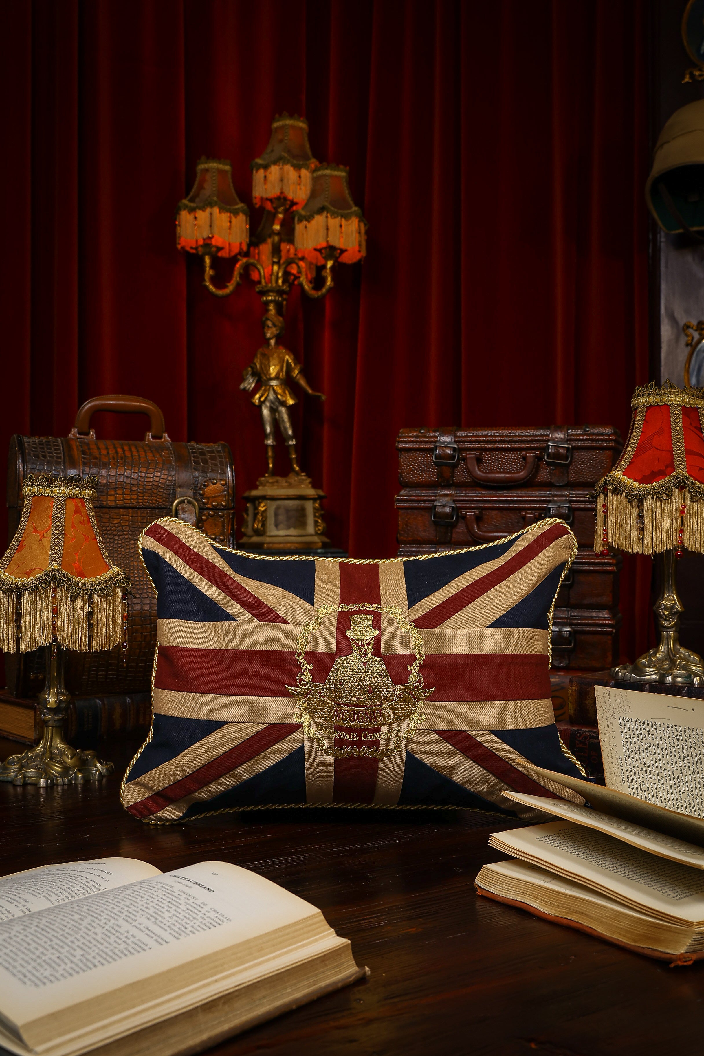 A rectangular cushion with a Union Jack design placed on a wooden surface, surrounded by a red-curtained window, a lamp, and a bookshelf in the background.