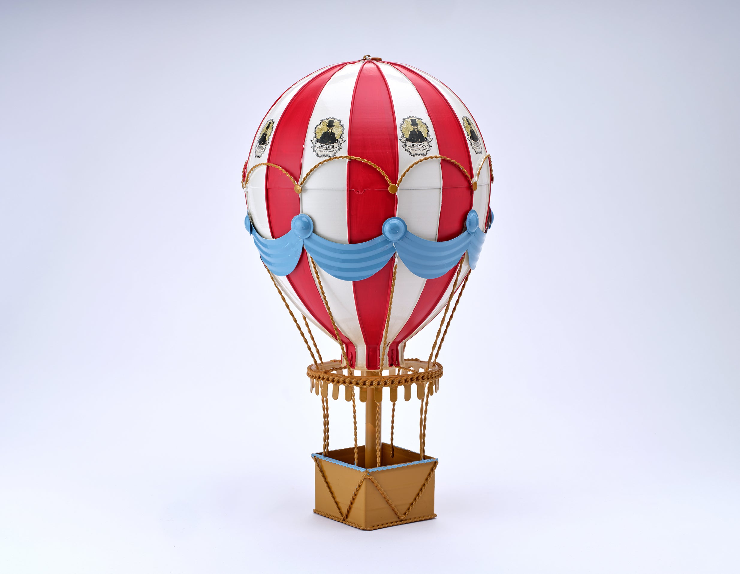 A miniature hot air balloon model with red and white vertical stripes, light blue draped accents, and a gold-colored basket, set against a clean light gray background.